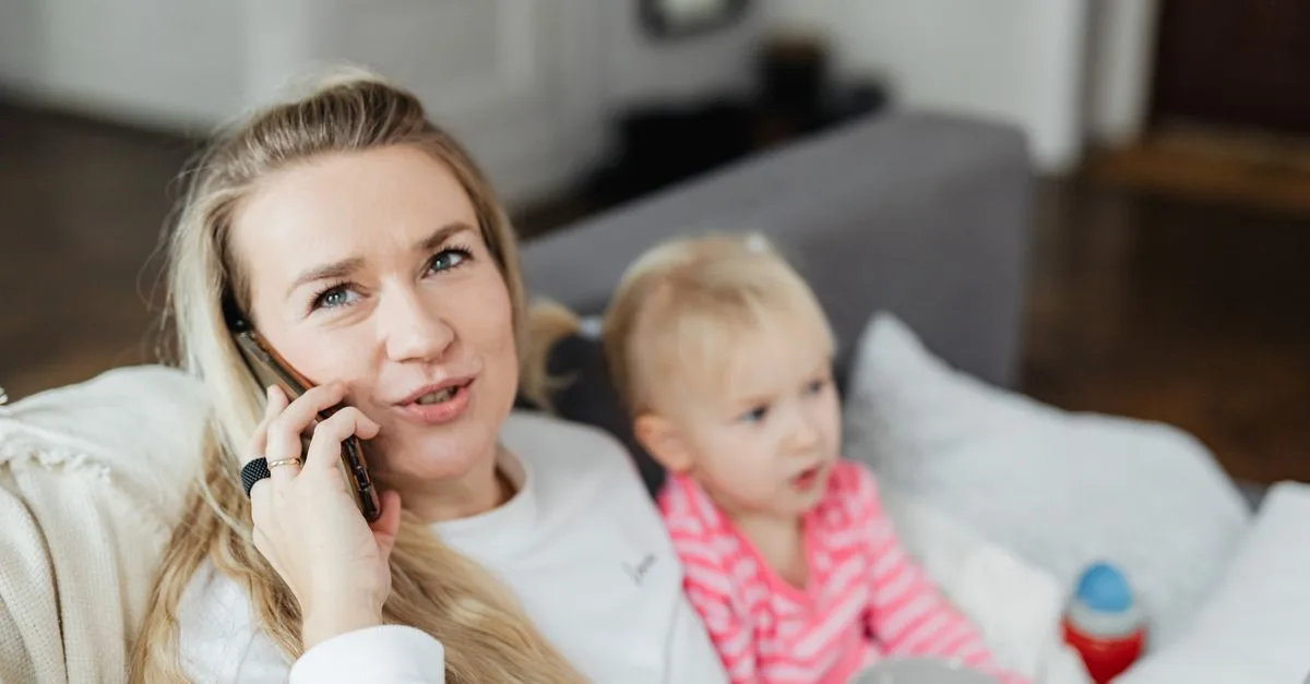 A parent talking on the phone at home with their young child beside them