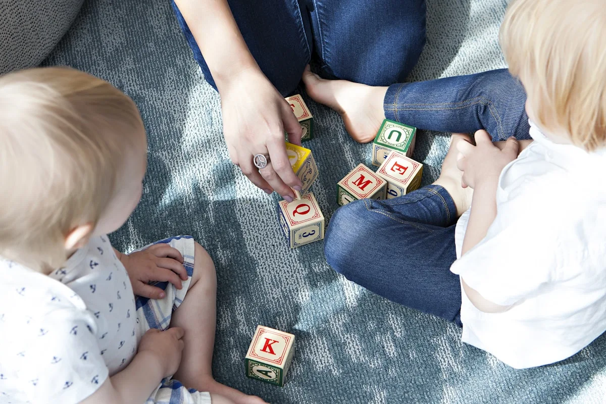 Two young children playing with alphabet blocks on the floor beside an adult